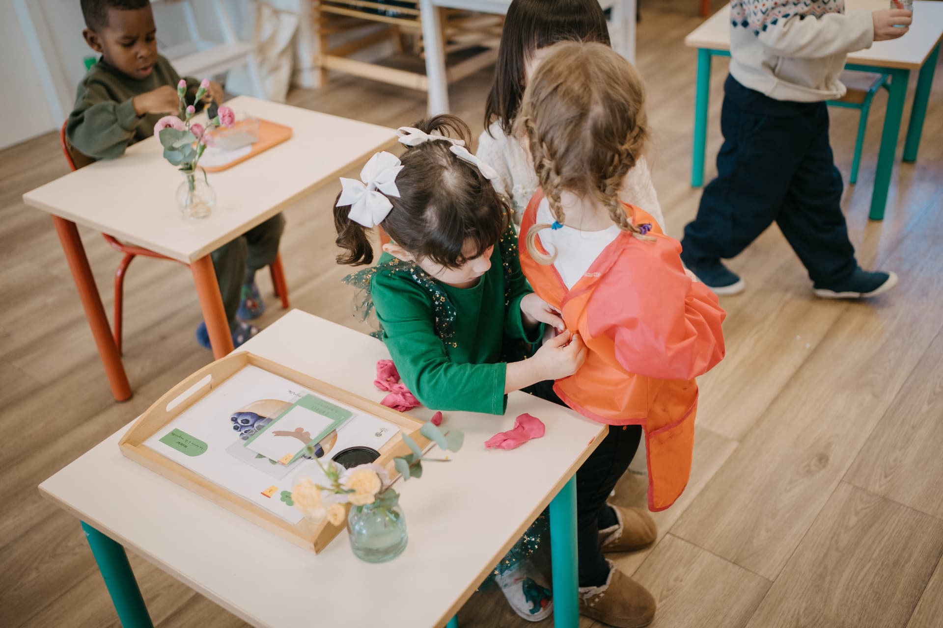 Une enfant aide une camarade à enfiler son tablier orange près d'une table de travail