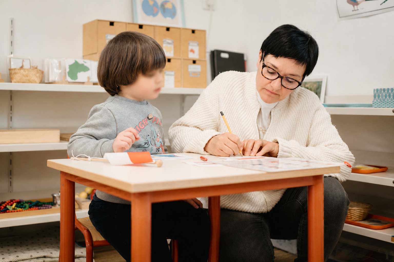 Corinne, éducatrice Montessori, écrit avec un enfant à une petite table de la classe