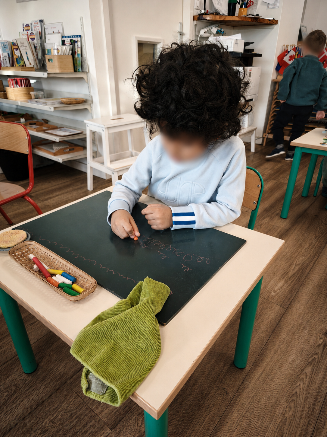 Un enfant trace des boucles à la craie sur une grande ardoise noire, assis à une petite table