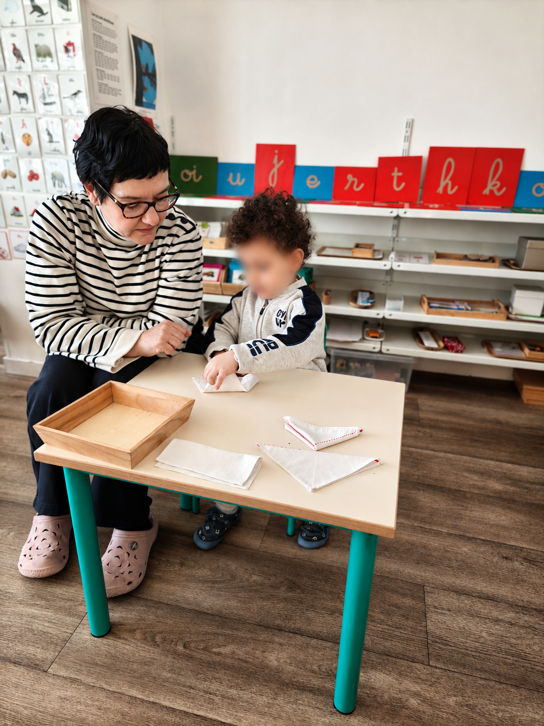 Une éducatrice, assise à côté d'un enfant, l'observe plier une serviette en tissu sans faire à sa place