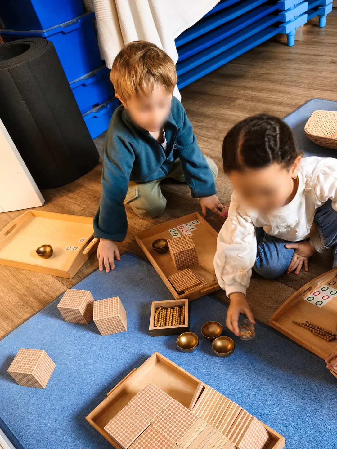 Deux enfants manipulent le matériel de perles dorées, cubes et plaquettes, installés sur un tapis bleu