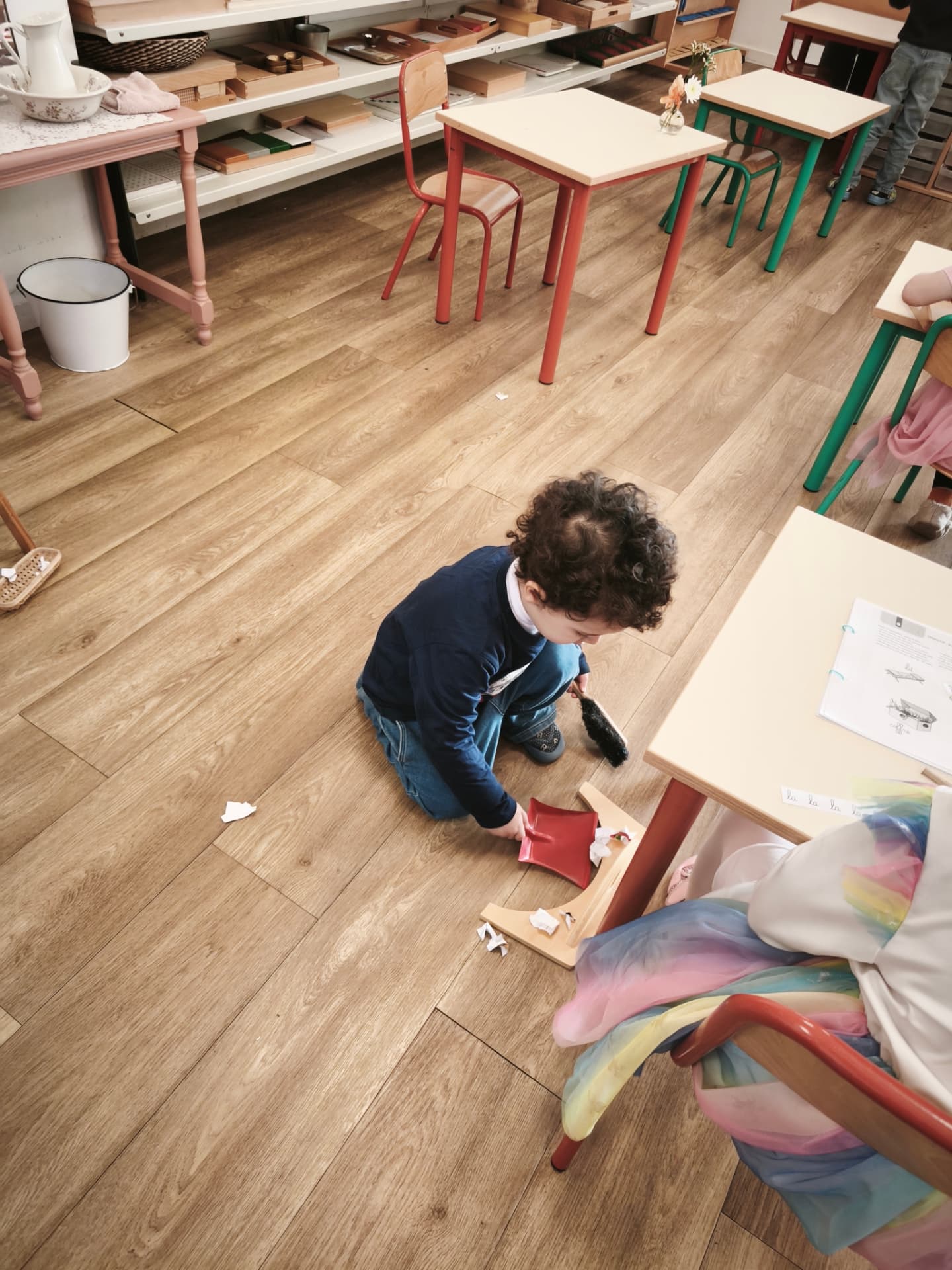 Un enfant agenouillé au sol ramasse des papiers avec une balayette et une pelle rouge dans la classe