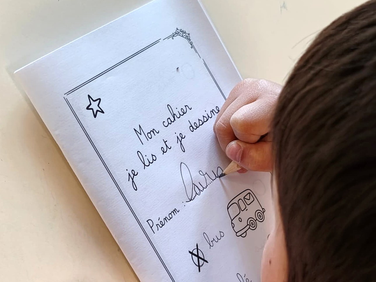 Gros plan sur un enfant qui écrit son prénom au crayon sur la couverture de son cahier « Mon cahier je lis et je dessine »