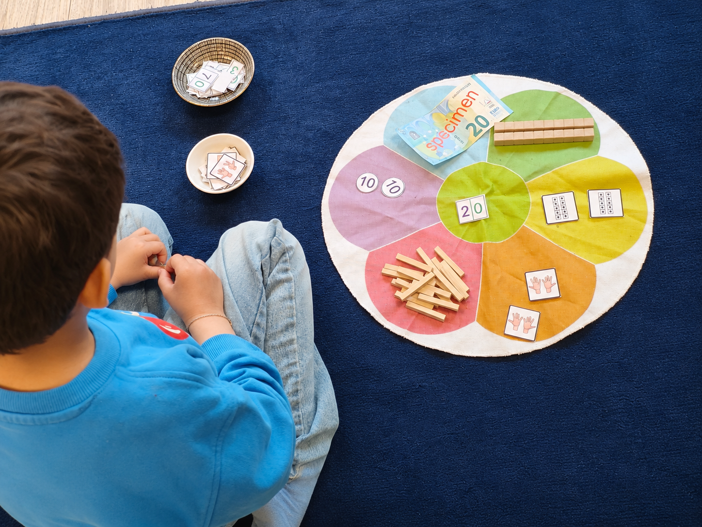 Un enfant assis au sol dispose des jetons, des barres et des cartes de nombres sur un tapis coloré de mathématiques