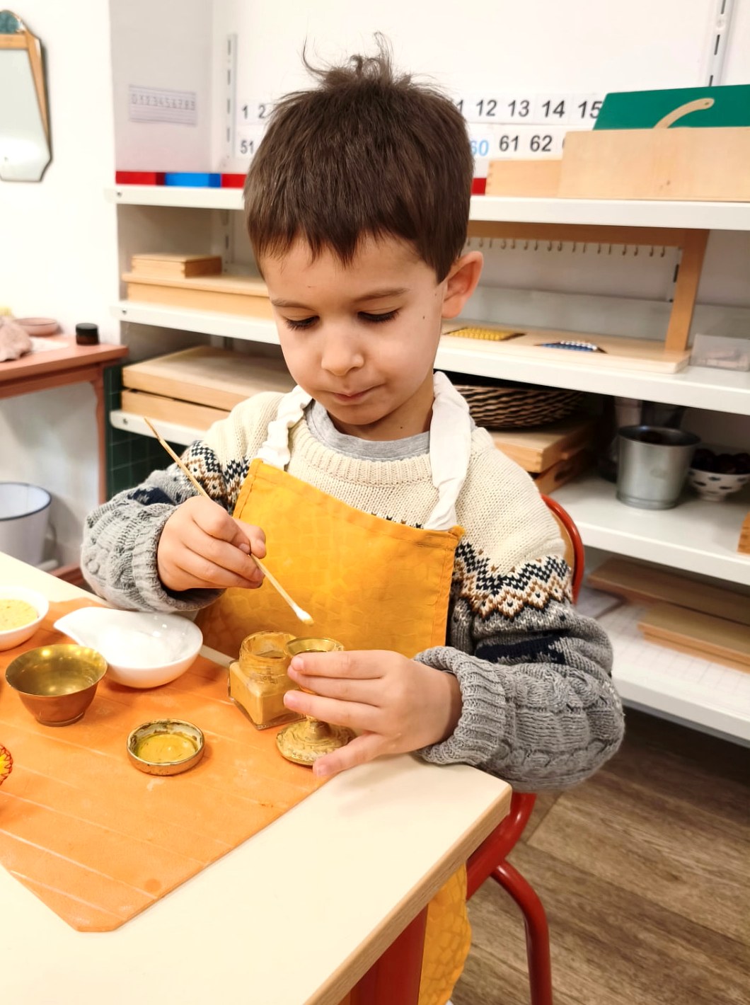 Un enfant en tablier jaune, concentré, applique de la pâte à polir sur un petit objet en laiton