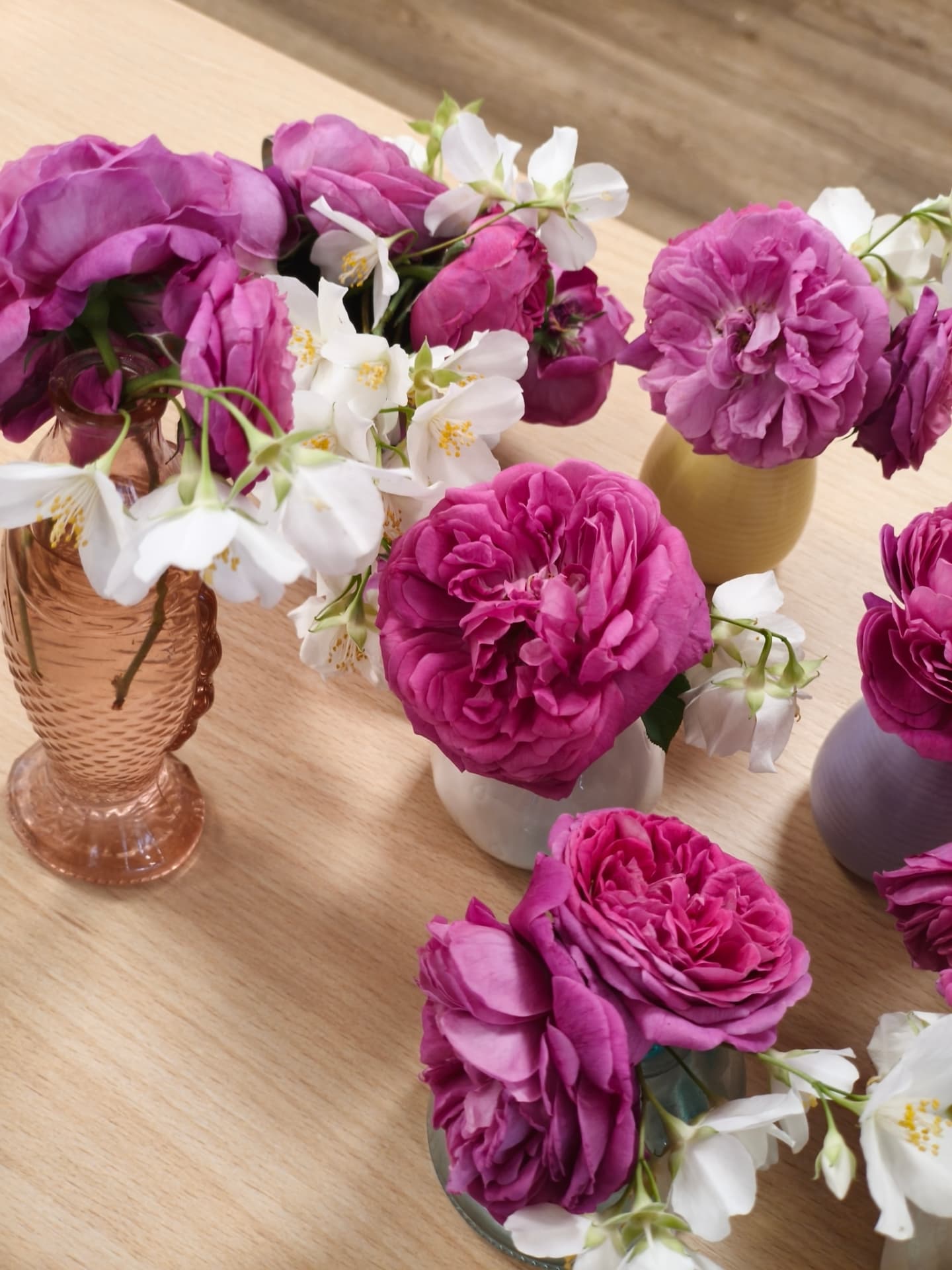 Plusieurs petits vases colorés garnis de roses et de fleurs blanches alignés sur une table en bois clair