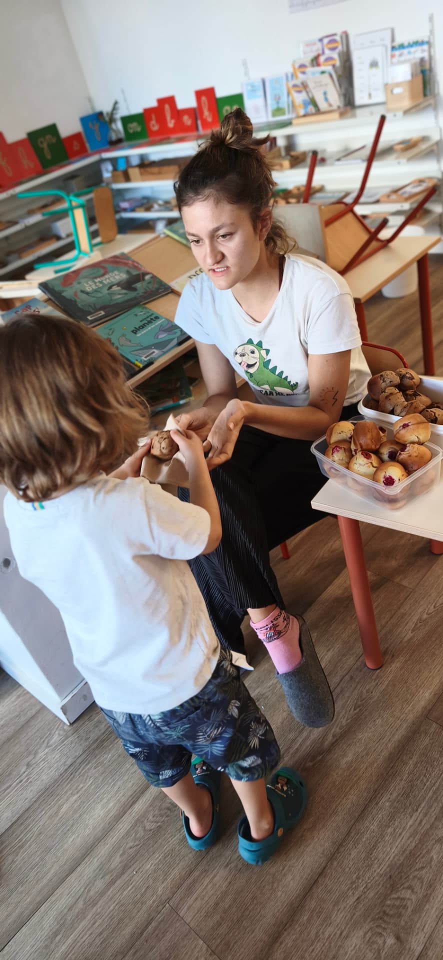 Une adulte tend un muffin à un enfant, une boîte de muffins posée sur la petite table à côté