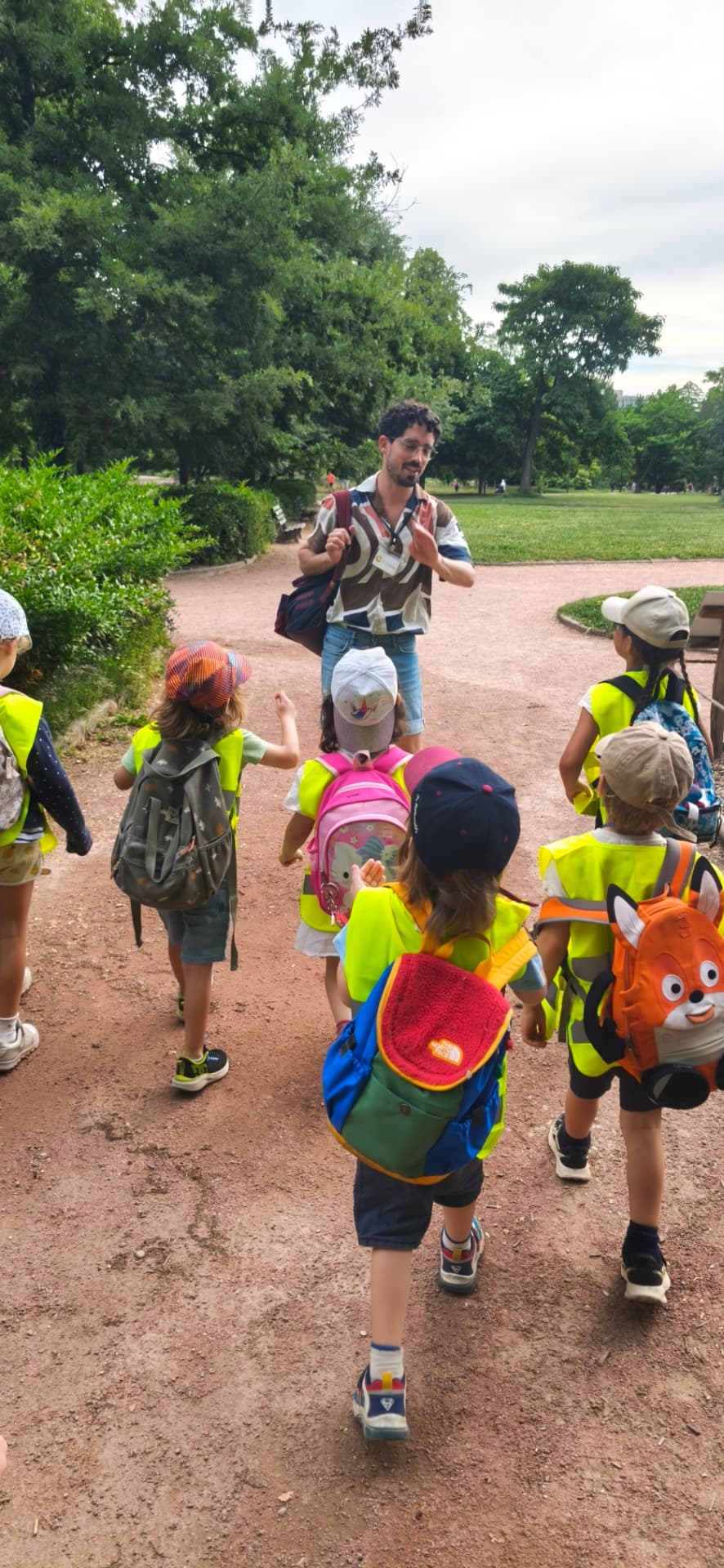 Un groupe d'enfants en gilets jaunes marche dans une allée du parc avec un éducateur