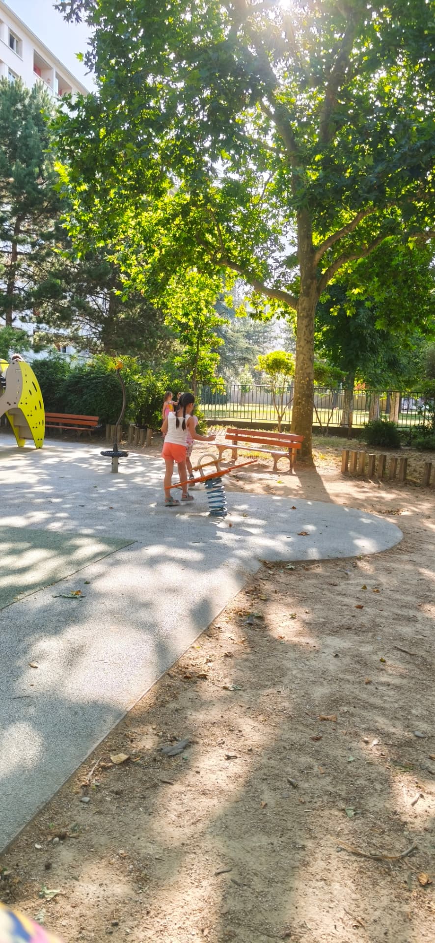 Deux enfants jouent autour du jeu à ressort du square, sous un grand arbre
